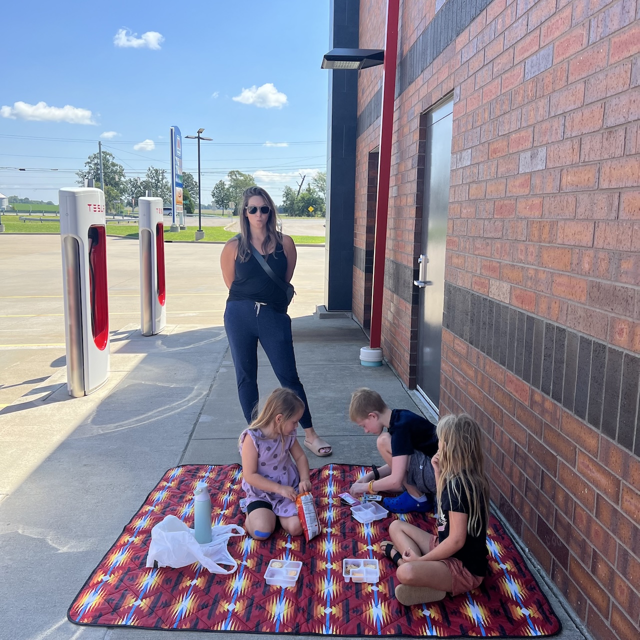 A picnic blanket in a parking lot with humans eating processed foods.